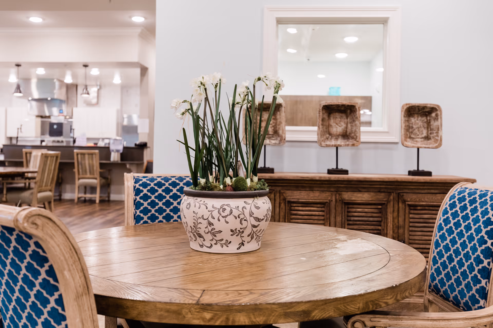 A round wooden table with a decorative white pot containing tall white flowers in the center. Surrounding the table are chairs with blue patterned upholstery. In the background, there is a wooden sideboard with three decorative square objects on stands and a large mirror above it. Further back, a kitchen area with hanging lights and wooden chairs is visible.