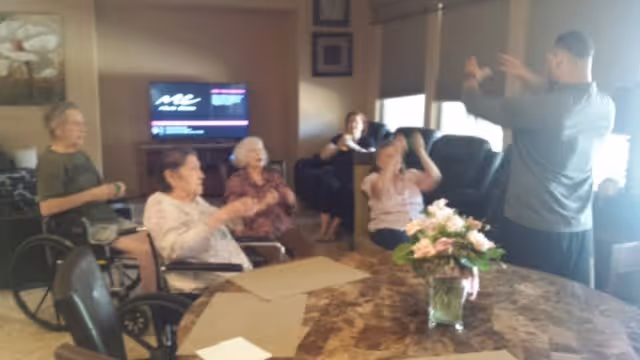 A group of elderly individuals, some seated in wheelchairs, participating in a group activity led by a standing instructor in a cozy living room setting with a TV mounted on the wall and a table with a flower arrangement in the foreground.