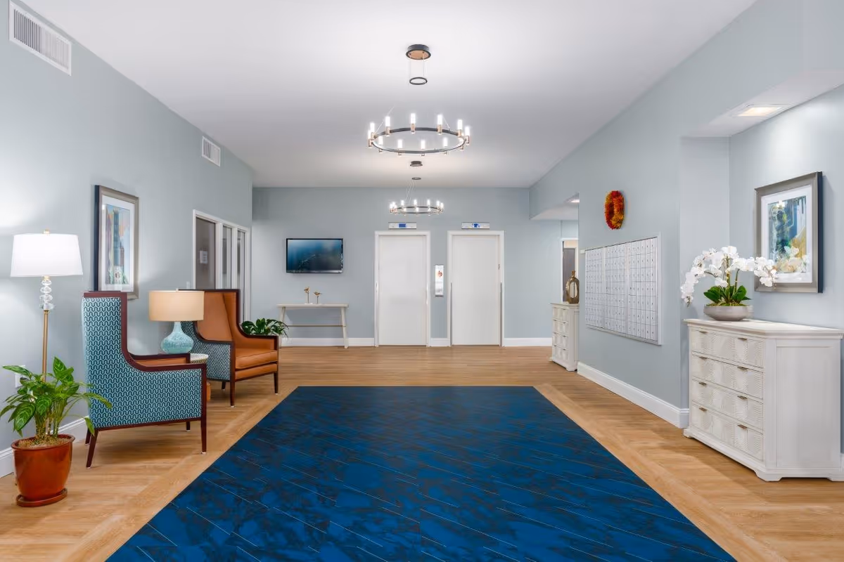 A spacious, well-lit lobby area with light blue walls and wooden flooring. There are two elevators at the far end, a blue patterned rug in the center, and seating with two armchairs and a side table with a lamp on the left. On the right side, there is a white chest of drawers with a potted orchid on top and a wall-mounted mailbox unit. The ceiling has modern circular light fixtures, and framed artwork is displayed on the walls.