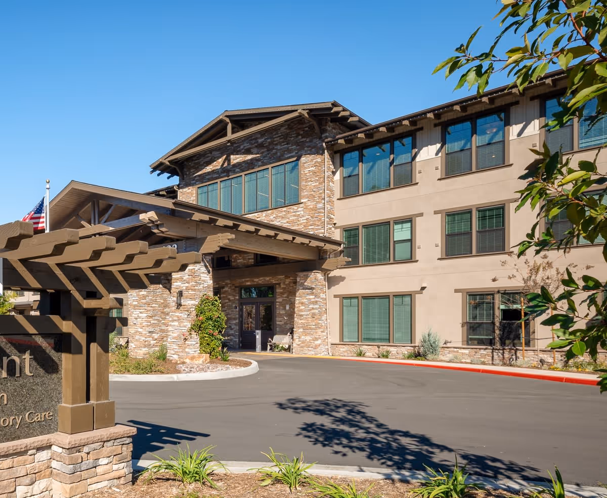 Exterior view of a senior living facility building with stone and beige walls, multiple windows, and a covered entrance. There is a paved driveway in front and some landscaping with plants and trees. An American flag is visible near the entrance under a clear blue sky.