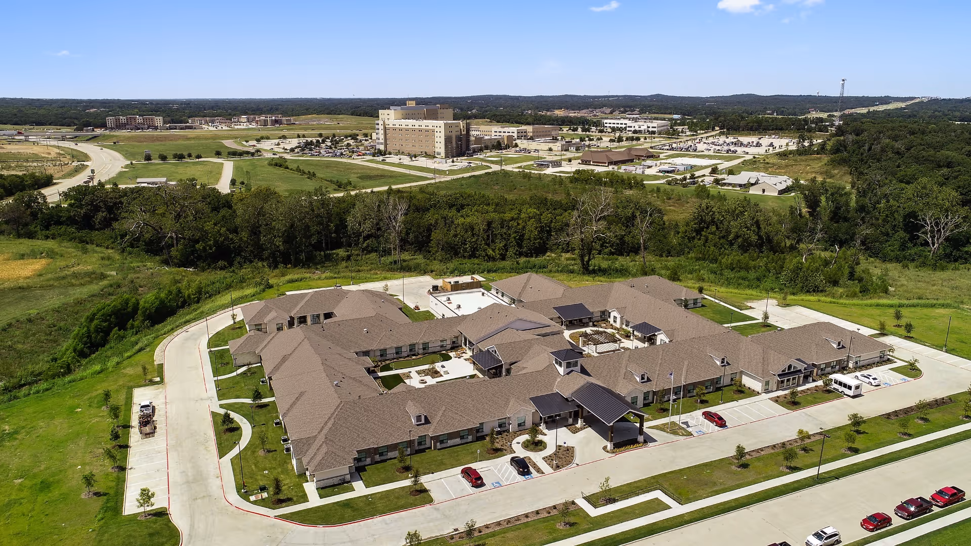 Aerial view of Celebration Senior Living of Denison, showing a large single-story building with multiple wings, surrounded by green lawns and parking areas. The facility is situated in a semi-rural area with trees and open fields nearby, and other buildings and roads are visible in the distance under a clear blue sky.