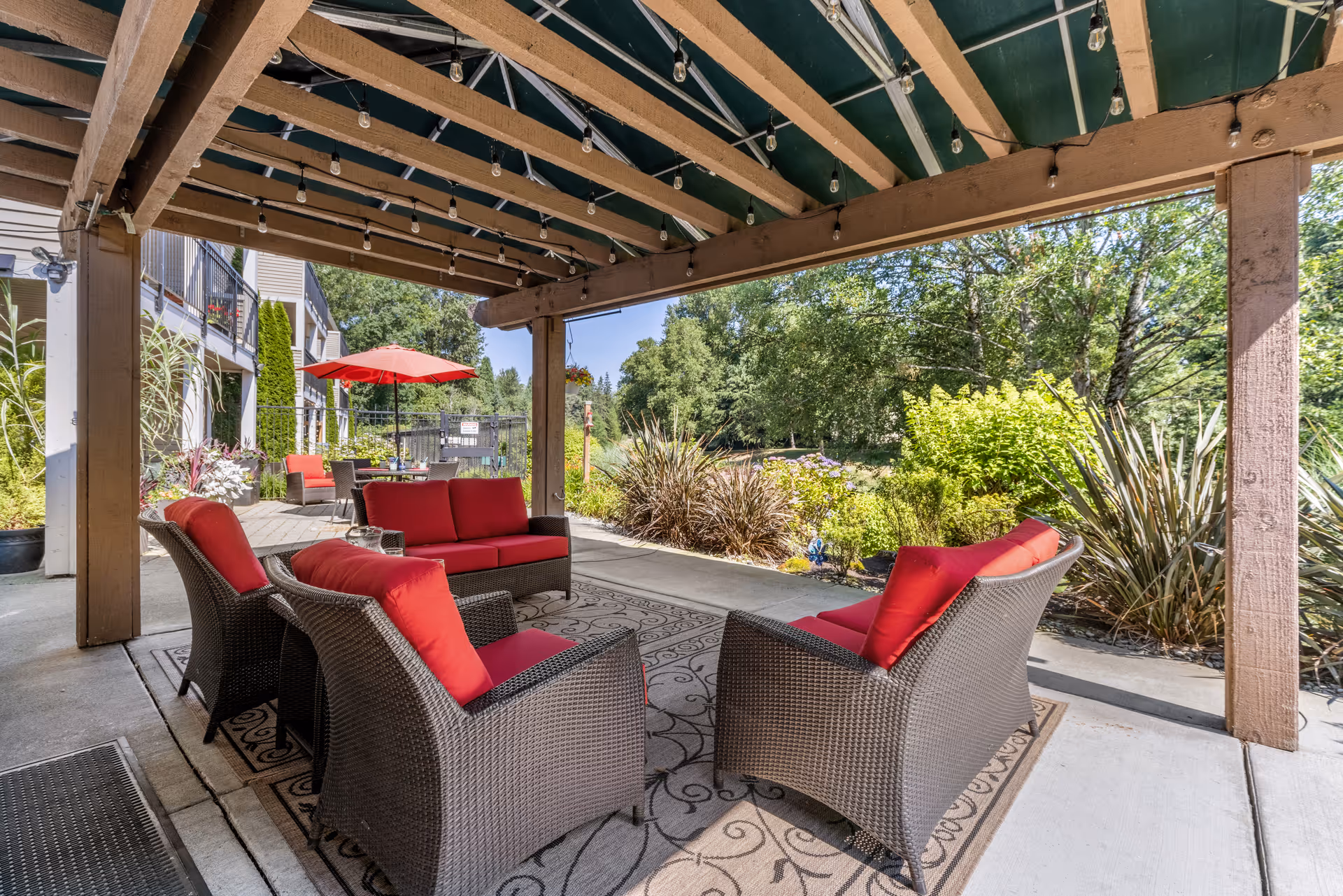 Outdoor covered patio area with wicker furniture featuring red cushions arranged around a patterned outdoor rug. There are string lights hanging from the wooden pergola overhead. In the background, there is a table with a red umbrella and additional seating, surrounded by greenery and trees.