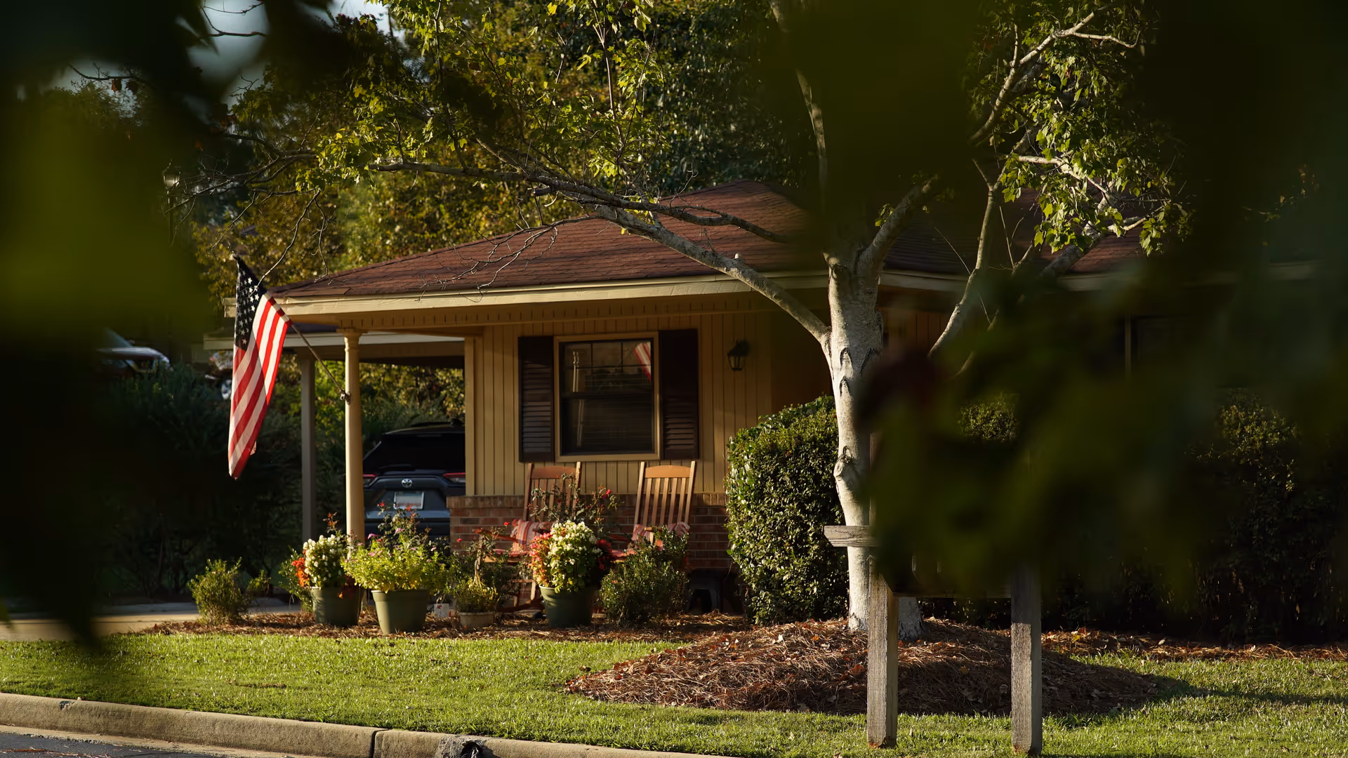 View of a small house with a covered porch featuring two wooden rocking chairs and several potted plants. An American flag is hanging from the porch, and a car is parked in the driveway. The scene is framed by tree branches and leaves in the foreground.