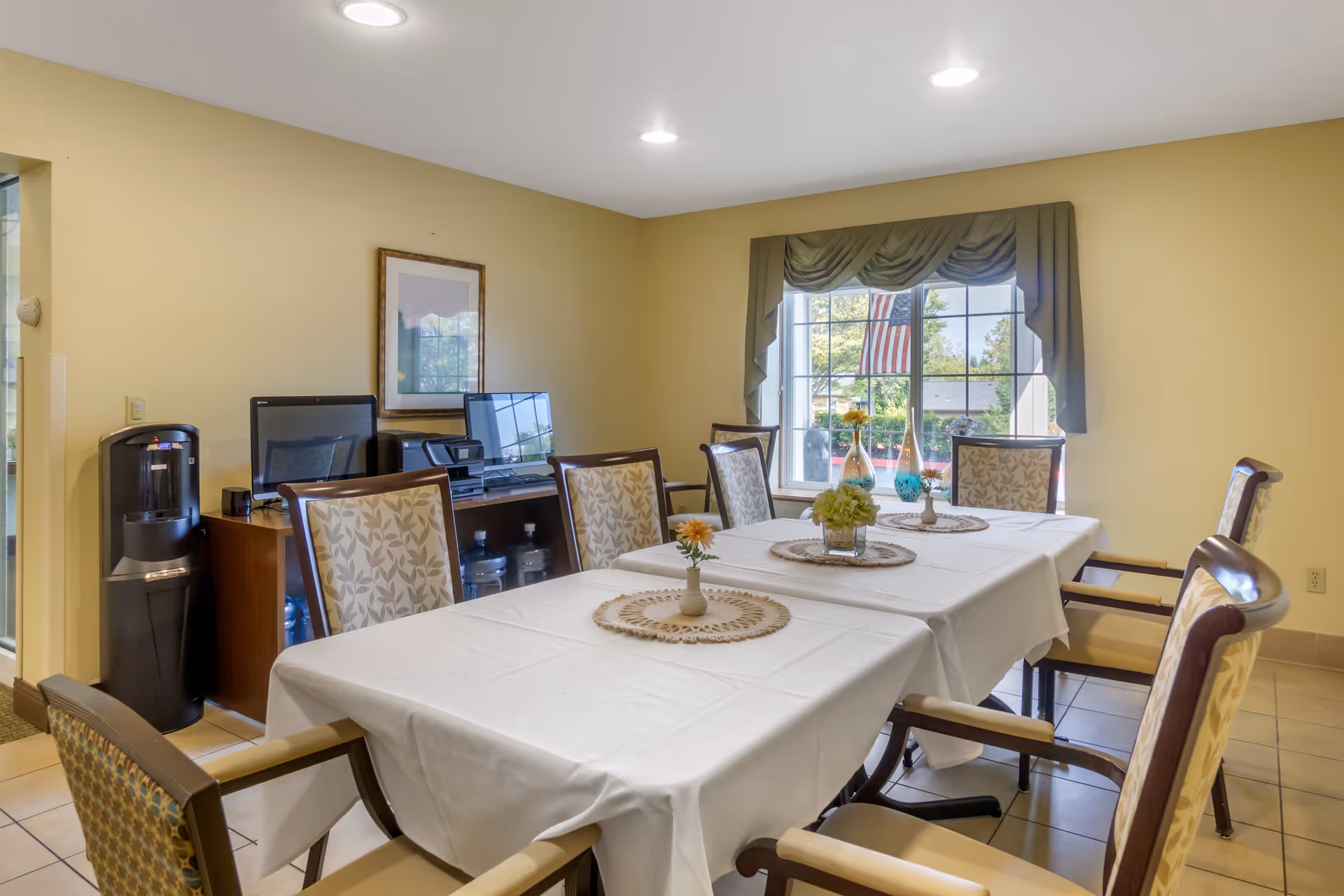 Bright dining room with a long table covered in a white tablecloth, floral centerpieces, and upholstered chairs in front of a window displaying an American flag.