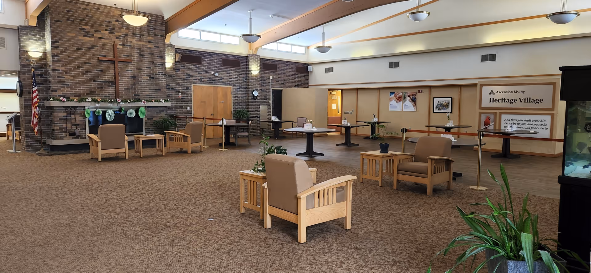 A spacious common area in a senior living facility with beige carpet and wooden furniture including armchairs and small tables. A large brick fireplace with a wooden cross above it is visible on the left side, decorated with flowers and green paper decorations. Several round tables with chairs are arranged in the background near a wall with signs that read 'Ascension Living Heritage Village.' The room is well-lit with ceiling lights and natural light from high windows. There are plants placed around the room and an American flag near the fireplace.