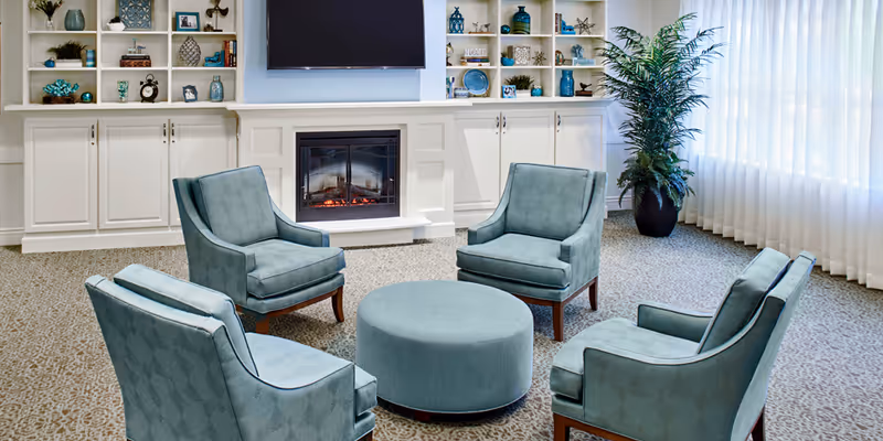 A cozy senior living common area with four teal upholstered armchairs arranged around a round teal ottoman. Behind the chairs is a white built-in cabinet with shelves displaying decorative items and a flat-screen TV mounted above an electric fireplace. A large potted plant is near a window with sheer white curtains allowing natural light to fill the room.