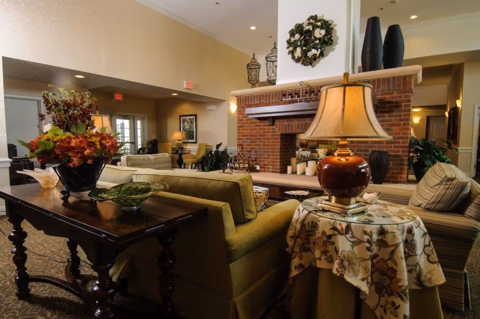 A cozy senior living facility common area with comfortable seating including sofas and armchairs arranged around a brick fireplace. There is a wooden table with a floral arrangement and a side table with a lamp covered by a floral tablecloth. The room is warmly lit with soft lighting and decorated with plants and wall art.