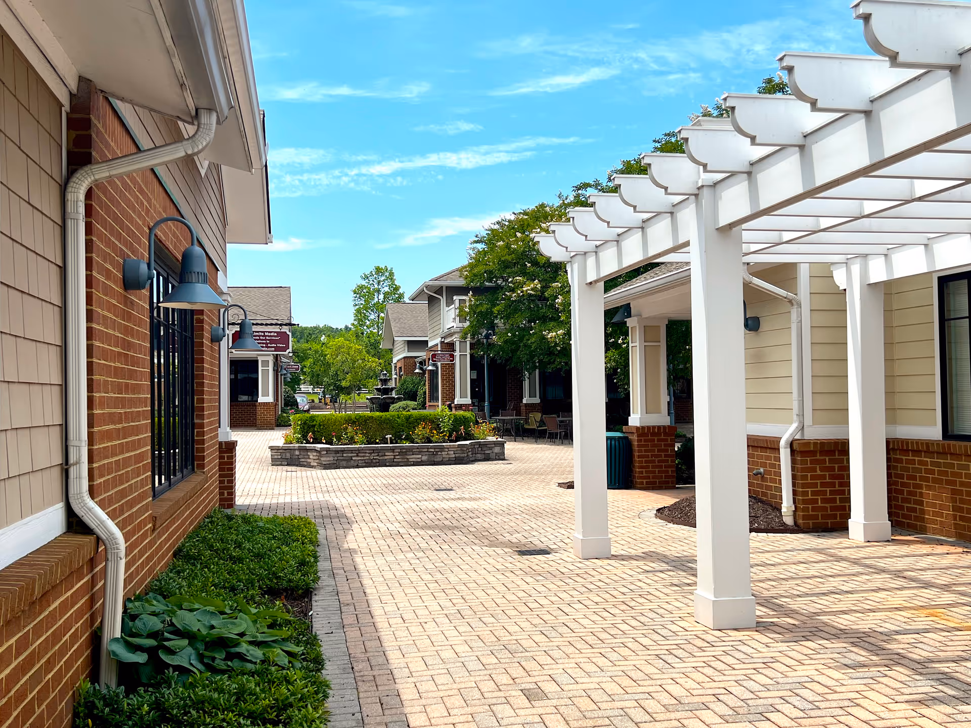 A sunny brick-paved courtyard with a white pergola, brick buildings, and landscaped planters and fountain.
