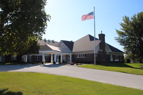 Exterior view of a single-story senior living facility building with a large American flag on a flagpole in front. The building has a covered entrance and is surrounded by green grass and trees under a clear blue sky.