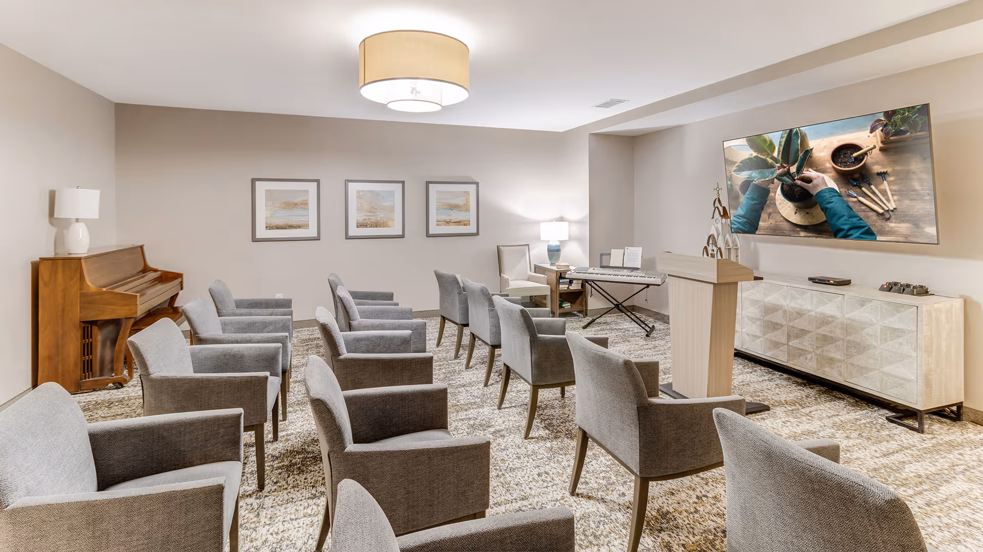 A well-lit room with multiple gray upholstered chairs arranged in rows facing a podium and a large wall-mounted TV screen displaying a person planting a small plant. The room also features a piano, a keyboard, a sideboard, three framed pictures on the wall, and two table lamps.