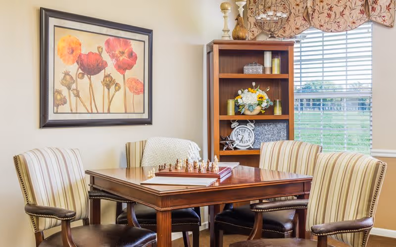 A cozy interior room with a wooden table set for a chess game, surrounded by four striped upholstered chairs. Behind the table is a wooden bookshelf with decorative items including candles, a clock, and a flower arrangement. A large window with blinds and a floral valance lets in natural light, and a framed floral painting hangs on the wall.