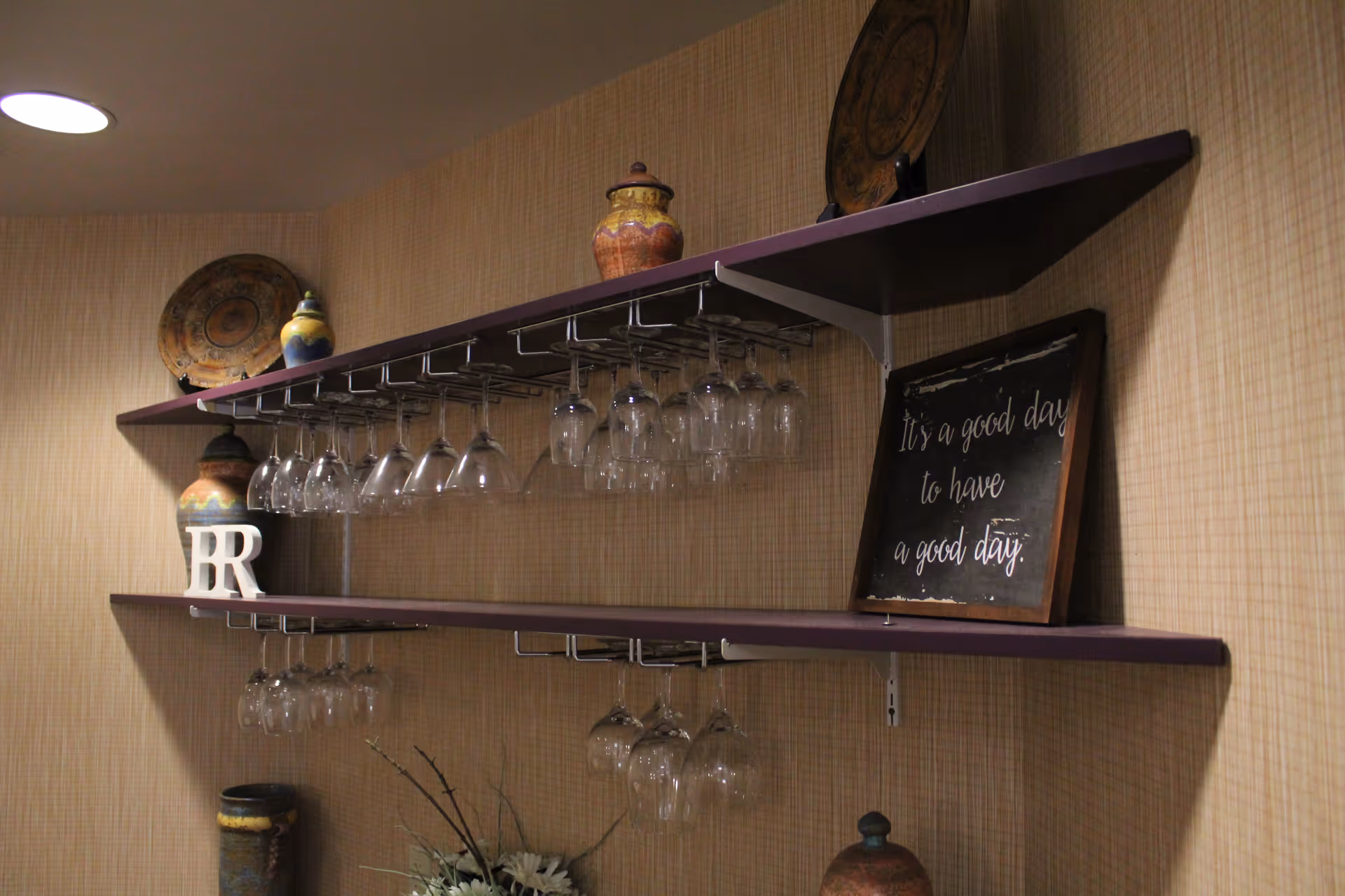 Two wall-mounted purple shelves holding decorative pottery, a framed sign that reads 'It's a good day to have a good day.', and several hanging wine glasses in a room with beige textured wallpaper.