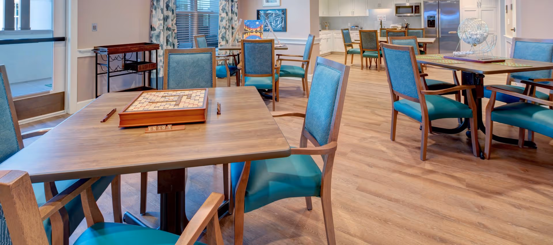 Communal dining/activity room with wooden tables and teal-upholstered chairs, a Scrabble board on the nearest table, and a kitchen area in the background.