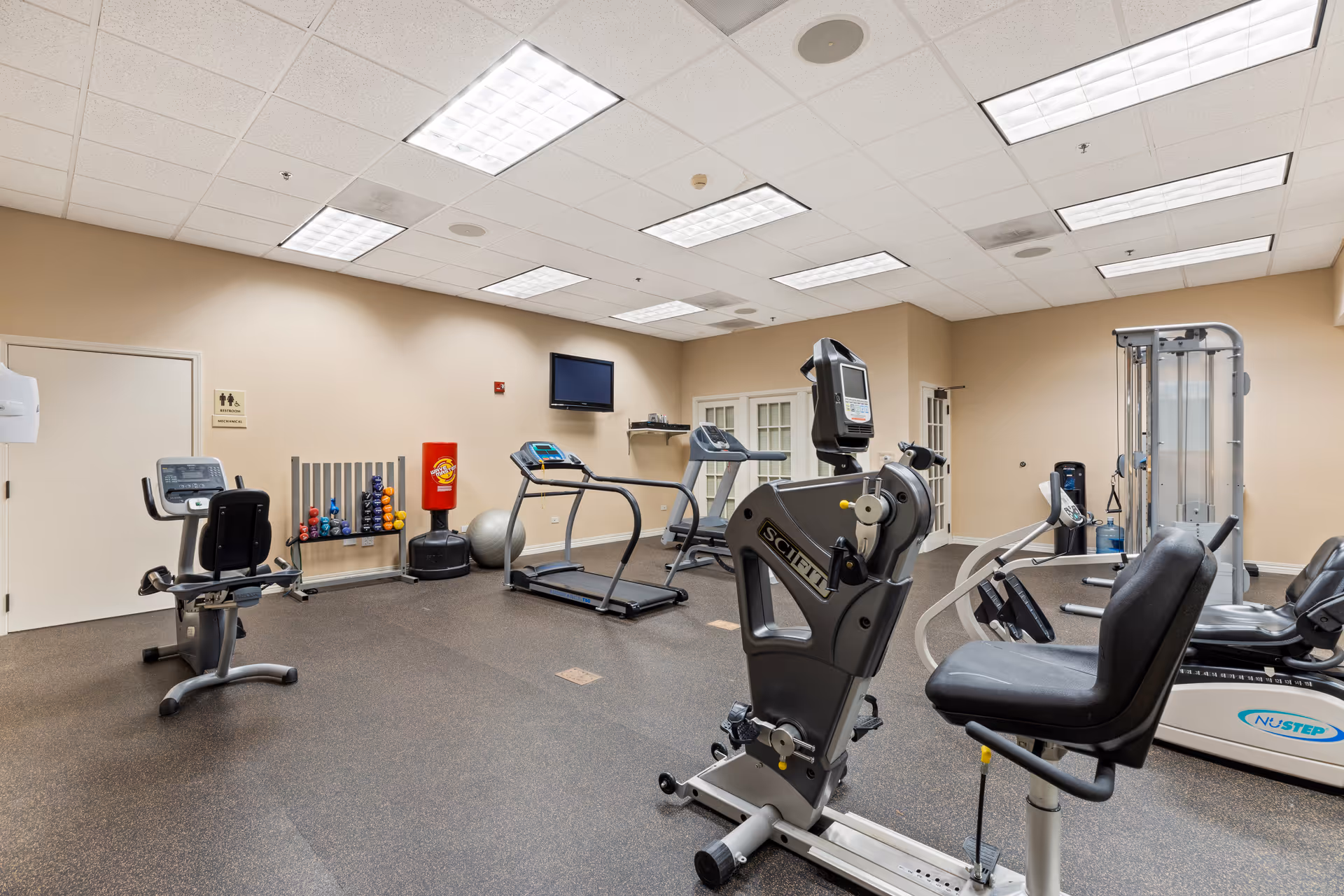 Interior view of a fitness room in a senior living facility with exercise equipment including stationary bikes, treadmills, a weight machine, and a rack of colorful dumbbells. The room has beige walls, a white drop ceiling with fluorescent lights, and a wall-mounted TV.