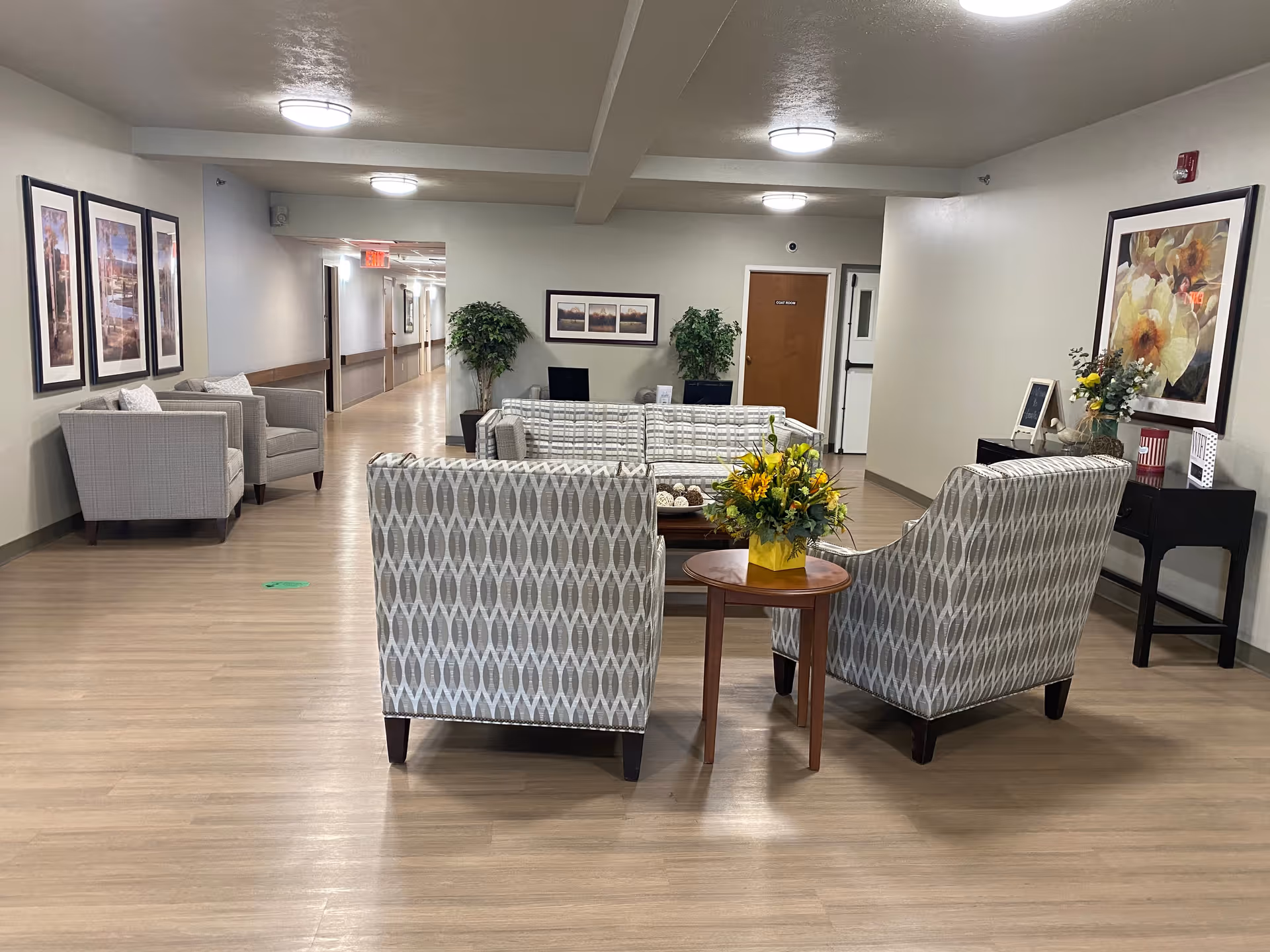 A well-lit common seating area in a senior living facility with patterned armchairs and a sofa arranged around a coffee table. There are framed pictures on the walls, potted plants, and a small table with a flower arrangement. A hallway extends from the seating area with doors along the sides.