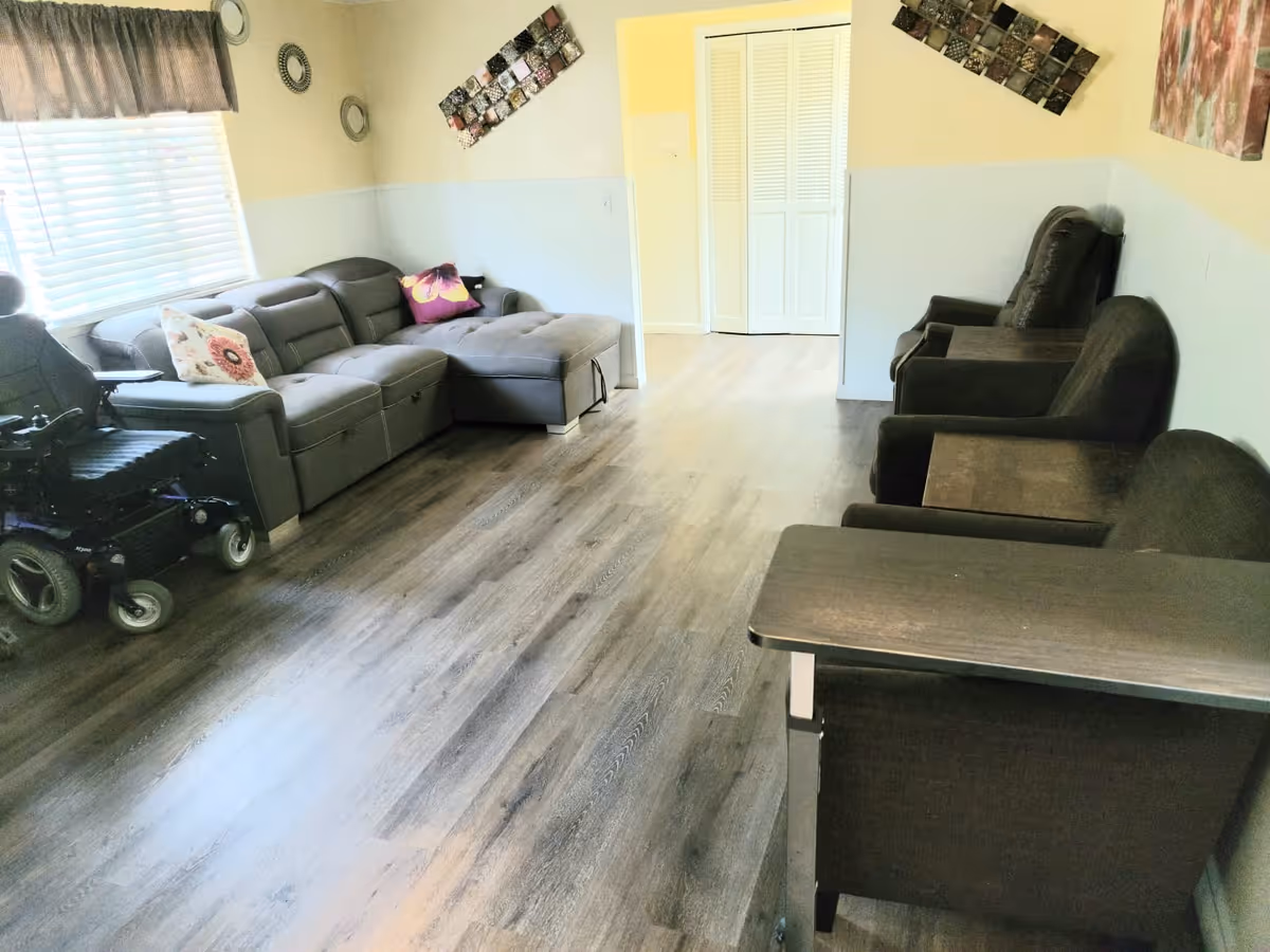 A living room with a gray sectional sofa adorned with decorative pillows, a power wheelchair next to the sofa, three dark armchairs with attached wooden side tables, light-colored walls with decorative wall hangings, and a window with blinds and a valance letting in natural light.