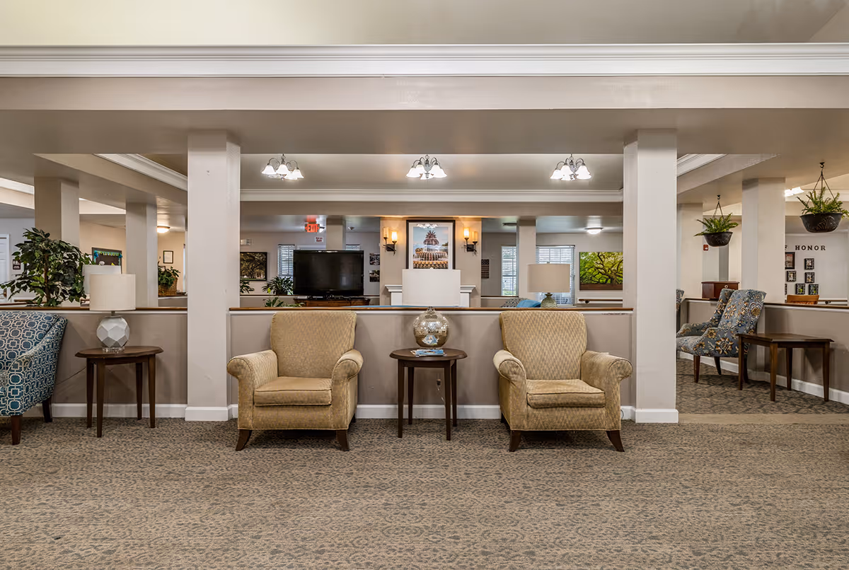 A spacious and well-lit common area with patterned carpet flooring, beige walls, and multiple seating options including two beige armchairs and a blue patterned chair. There are small wooden side tables with lamps and decorative items, hanging plants, and framed artwork on the walls. The area has ceiling lights and a TV visible in the background.