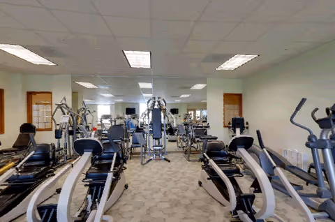 A senior living community fitness room with various exercise equipment including recumbent bikes, elliptical machines, and weight machines. The room has a carpeted floor, white ceiling with fluorescent lights, and mirrors on the walls reflecting the equipment.