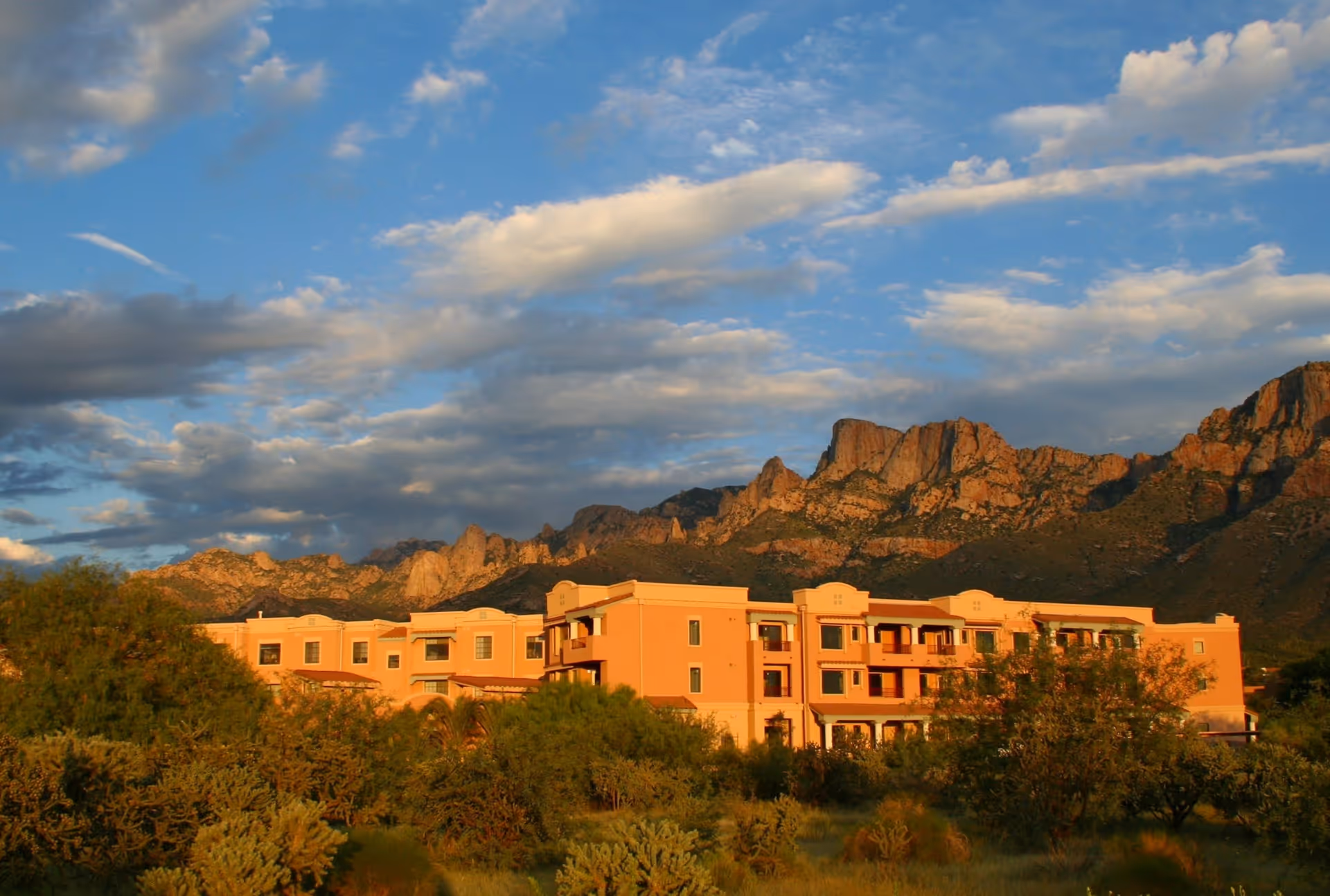 A large, peach-colored building set against a backdrop of rugged mountains under a partly cloudy blue sky, surrounded by desert vegetation.