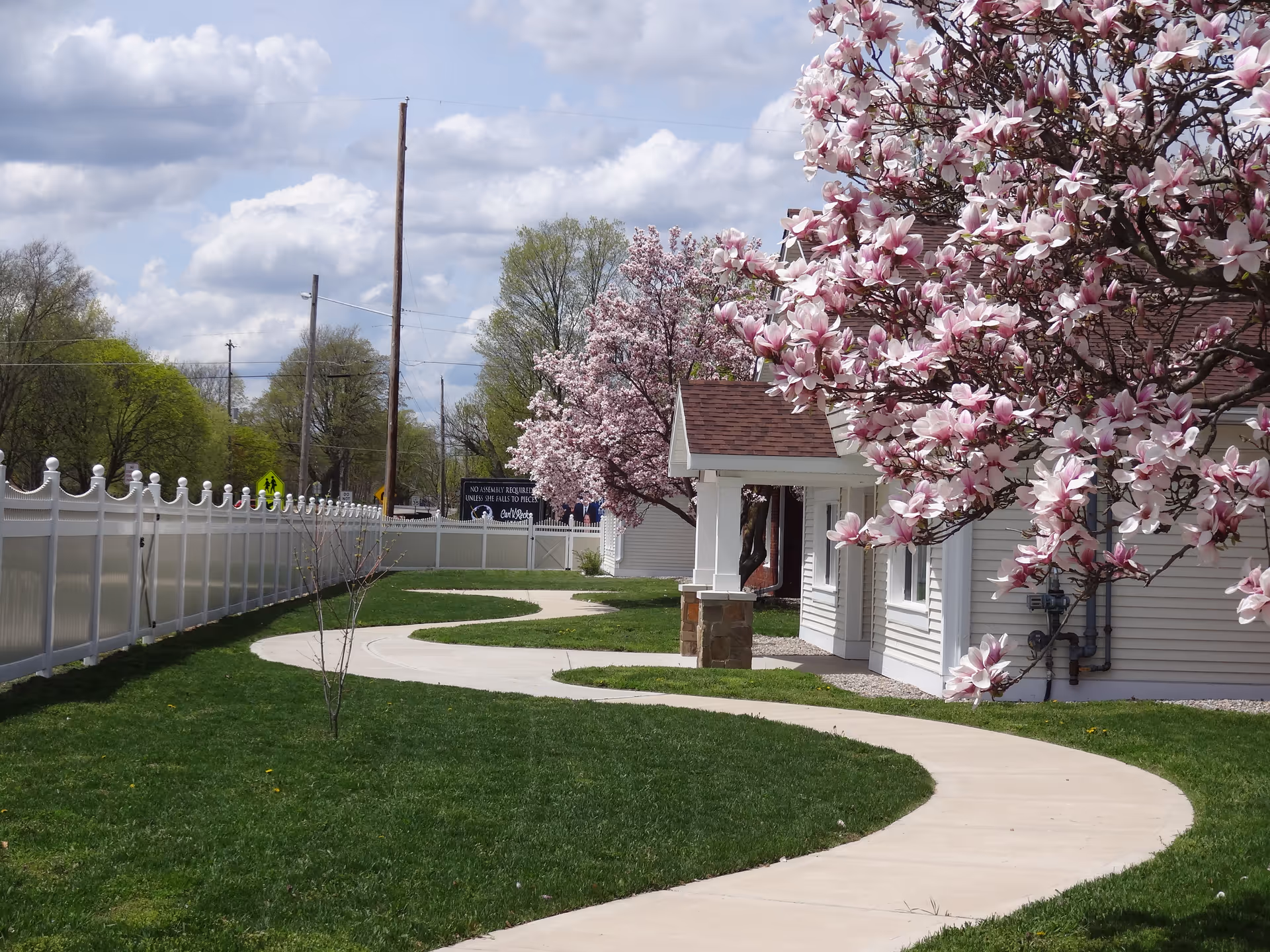 A curving concrete path winds through a grassy yard past a light-colored building and blooming pink magnolia trees under a partly cloudy sky.