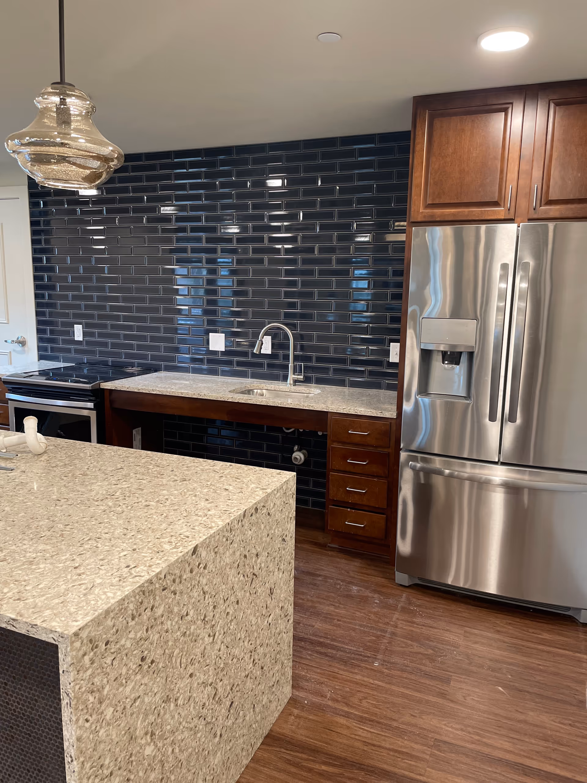 Modern kitchen with a large island countertop in the foreground, a stainless steel refrigerator with a water dispenser on the right, dark wood cabinets, a sink with a chrome faucet, and a black subway tile backsplash. The floor is wooden and there is a decorative hanging light fixture above the island.