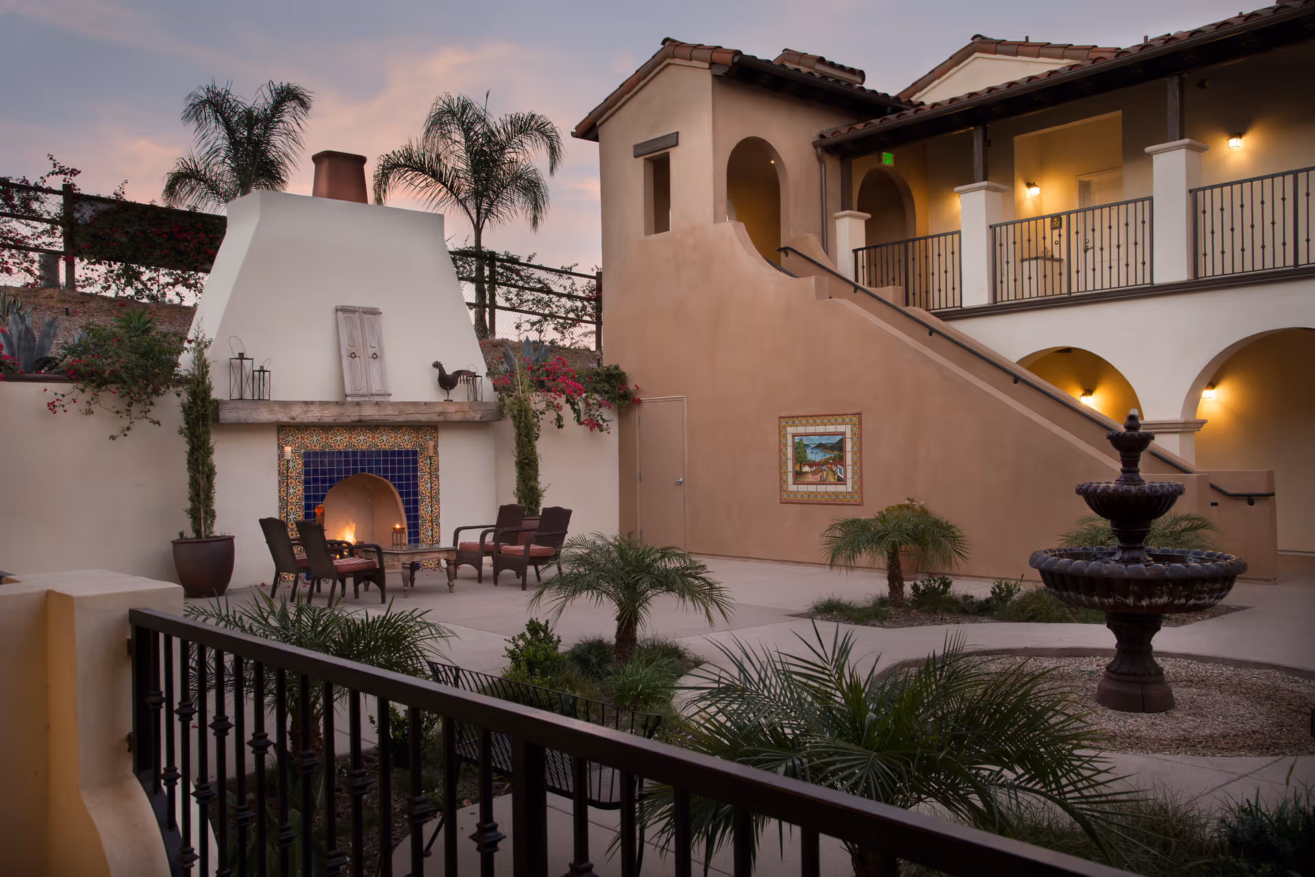 Outdoor courtyard area at Casa Aldea at University City Village featuring a large white fireplace with blue tile accents, surrounded by chairs and a table. The courtyard includes palm trees, potted plants, a multi-tiered fountain, and a two-story building with arches and a staircase. The scene is captured at dusk with warm lighting.