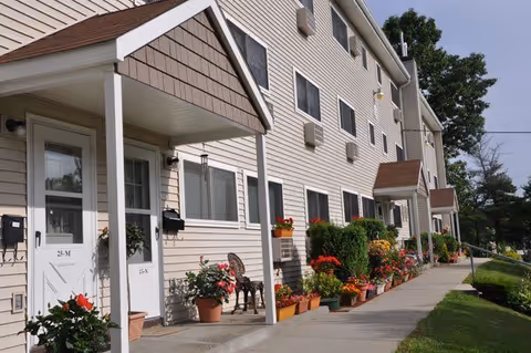 Exterior view of a multi-unit residential building with beige siding and brown shingled roofs over the entrances. The sidewalk is lined with numerous potted plants and flowers, and there is a green lawn on the right side. The sky is clear with some trees visible in the background.