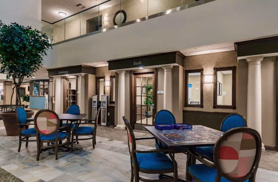 Interior view of a common area in a retirement community featuring tables and chairs with blue cushions, a partially completed jigsaw puzzle on one table, potted plants, and doors labeled 'Conference' and 'Bank' with classical columns framing the entrances.