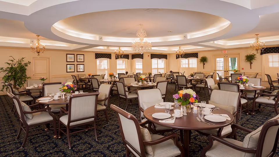 A spacious and elegant dining room with multiple round tables set with white plates, cups, glasses, and silverware. Each table has a colorful floral centerpiece. The room features a patterned carpet, beige walls with framed artwork, large windows with valances, and ornate chandeliers hanging from a decorative ceiling.