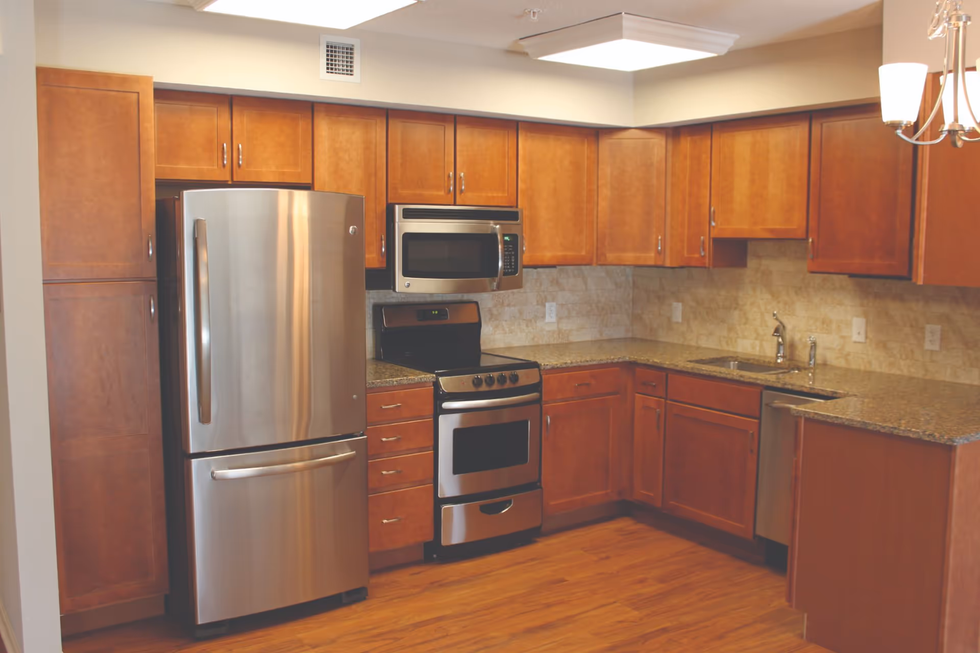 Modern kitchen with wooden cabinets, stainless steel refrigerator, oven with stove, microwave, dishwasher, granite countertops, and a double sink under a chandelier light fixture.
