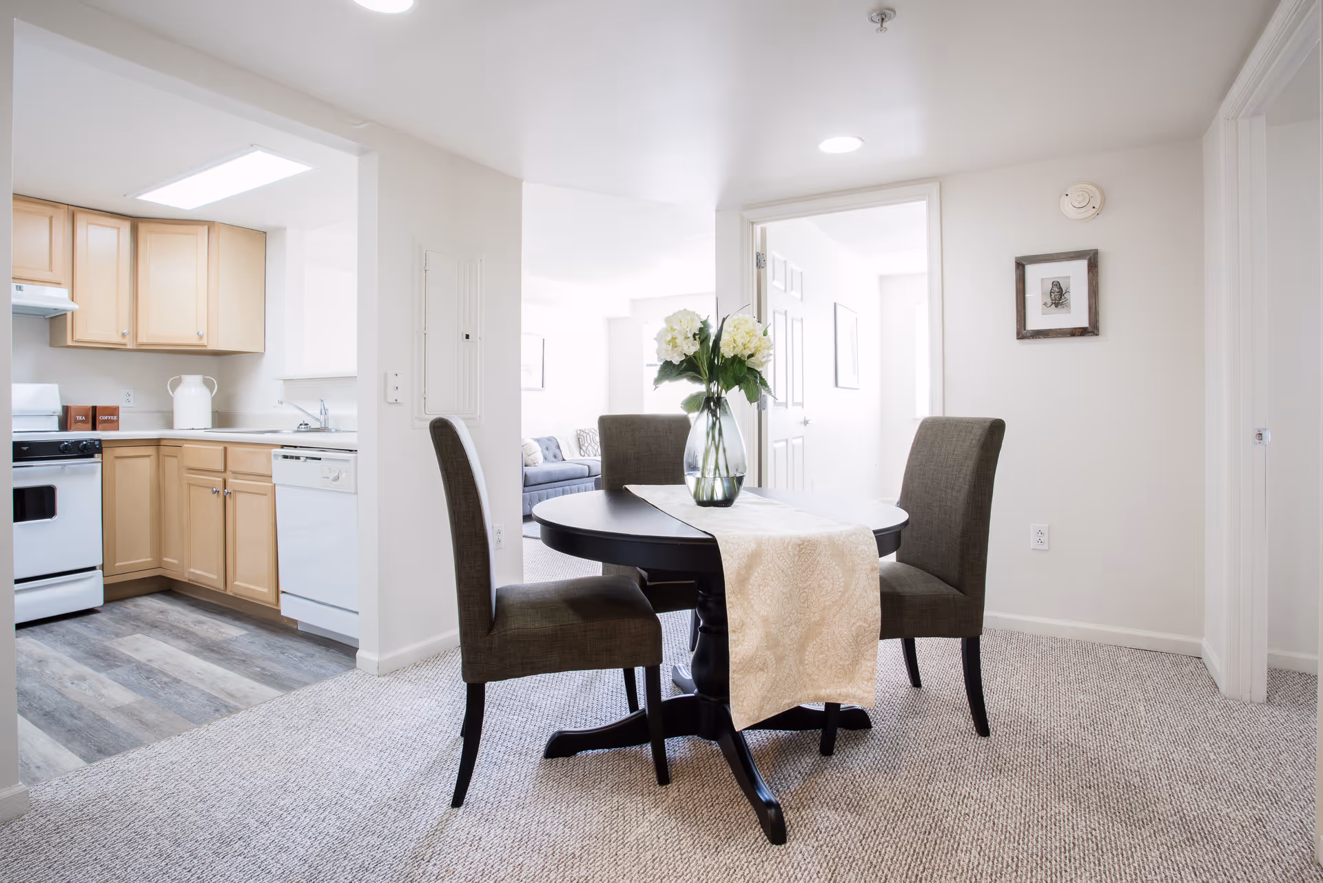 A bright dining area with a round black table covered by a beige table runner and a vase with white flowers in the center. Three upholstered chairs surround the table. To the left is a kitchen with light wood cabinets, a white stove, dishwasher, and a sink. The floor in the kitchen is wood-style vinyl, while the dining area has light carpet. In the background, there is a doorway leading to another room with a couch and framed pictures on the walls.