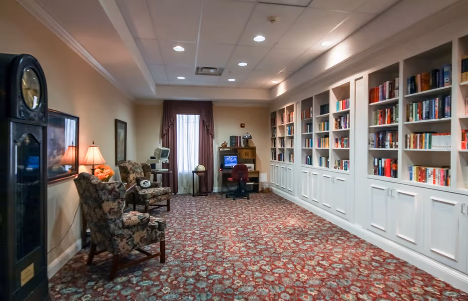 A cozy library or reading room with floral patterned armchairs, a tall grandfather clock, a table lamp, and a long wall of built-in white bookshelves filled with books. At the far end, there is a desk with a computer and a red office chair, and a window with curtains letting in natural light.