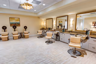 A spacious and well-lit salon area with beige styling chairs in front of large mirrors mounted on a wall with cabinets below. The room has light-colored flooring, a chandelier, and decorative artwork on the walls.
