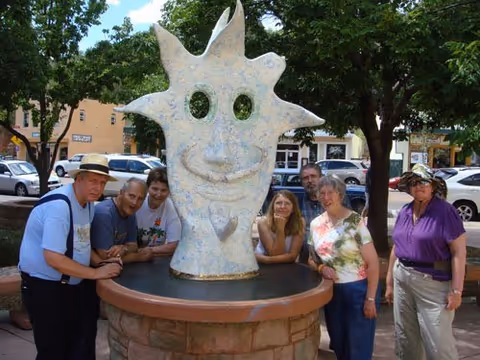 A group of six seniors standing around a circular stone fountain with a whimsical sun-shaped sculpture in the center, outdoors in a shaded area with trees and parked cars in the background.