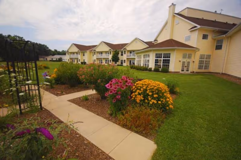 Yellow senior living building with a landscaped garden, colorful flowers and a paved walkway in the foreground.
