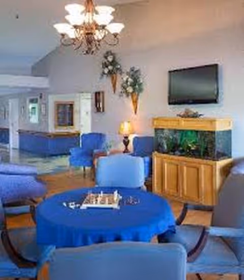 Senior living lounge with blue seating, a round table, chandelier, and aquarium cabinet.