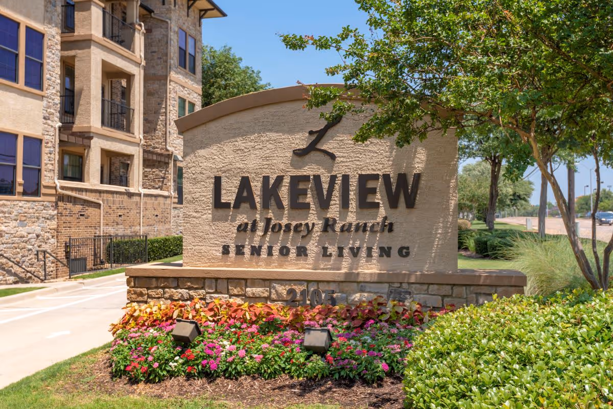 Stone sign for Lakeview at Josey Ranch Senior Living with colorful flowers and greenery in front, and a multi-story stone building in the background under a clear blue sky.