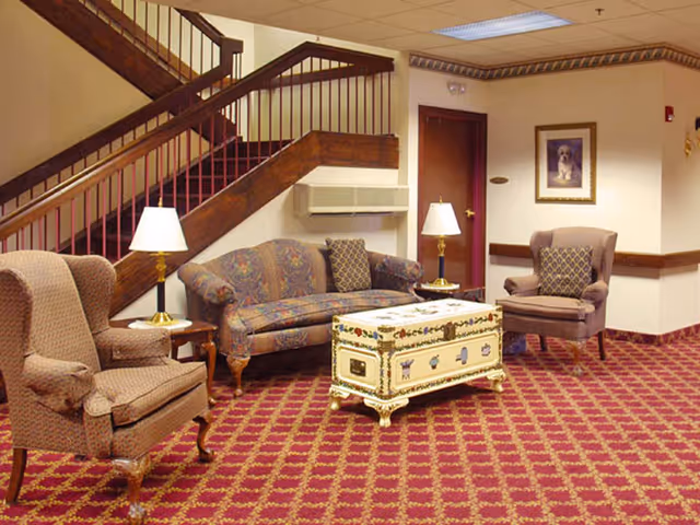 A cozy sitting area in a senior living facility with two upholstered armchairs and a patterned sofa arranged around a decorative trunk used as a coffee table. The room features a red and gold patterned carpet, wooden staircase with dark wood railing, two table lamps on side tables, and a framed picture on the wall.