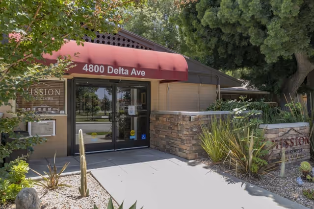 Entrance to Mission Care Center with a red awning displaying the address 4800 Delta Ave. The building has glass double doors and is surrounded by plants and trees.