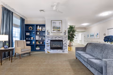 Bright assisted-living common room with a central fireplace flanked by blue built-in shelves, a gray sofa and armchair, and a hallway to the right.