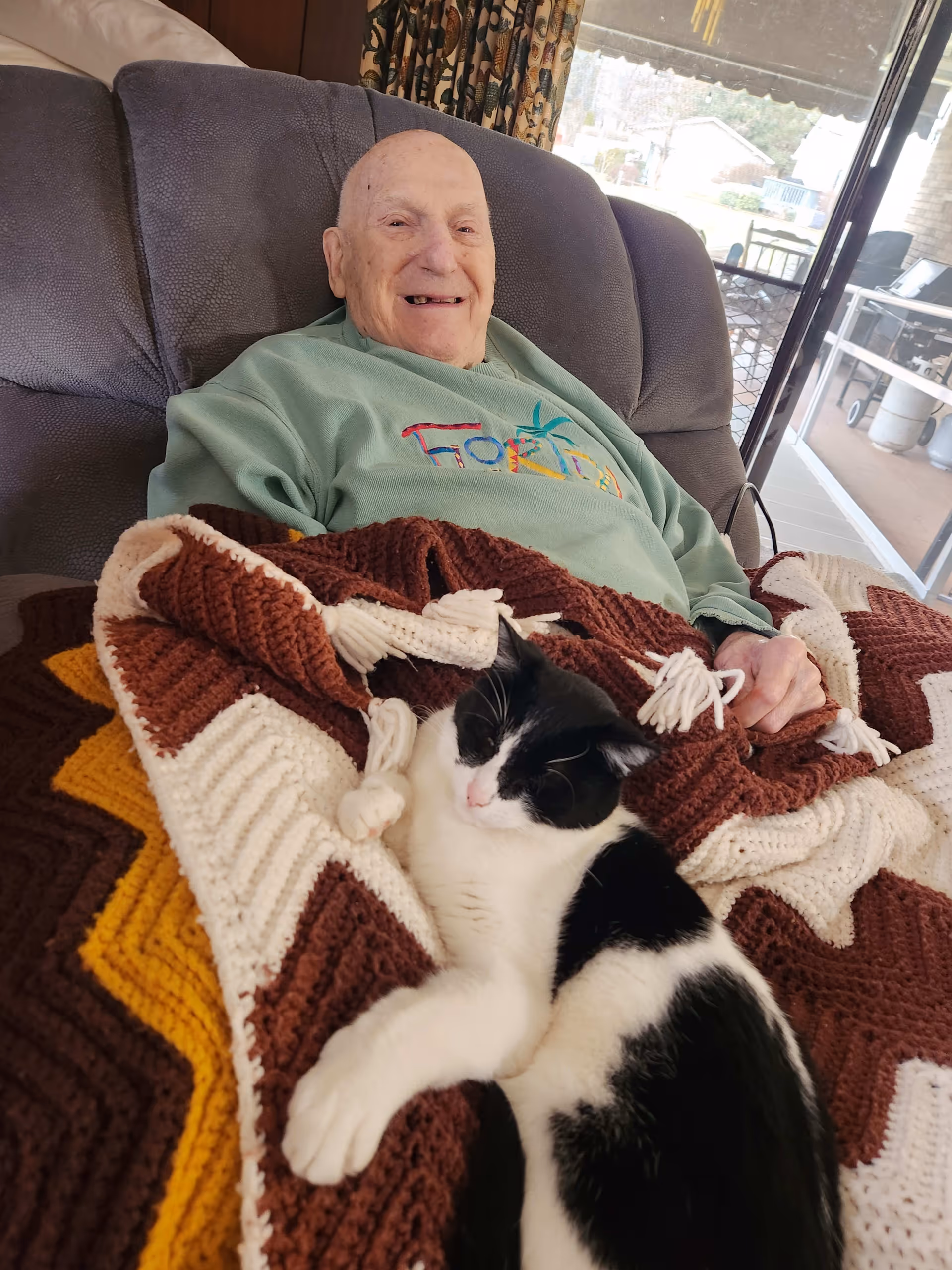 An elderly man sitting comfortably on a gray recliner chair with a crocheted blanket in shades of brown, white, and yellow draped over his lap. A black and white cat is resting on the blanket. The man is smiling and wearing a green sweatshirt. Behind him is a window showing an outdoor patio area.