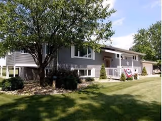 A single-story residential building with gray siding and white trim, featuring a front porch decorated with patriotic bunting. The house is surrounded by a well-maintained lawn, bushes, and a large tree providing shade.