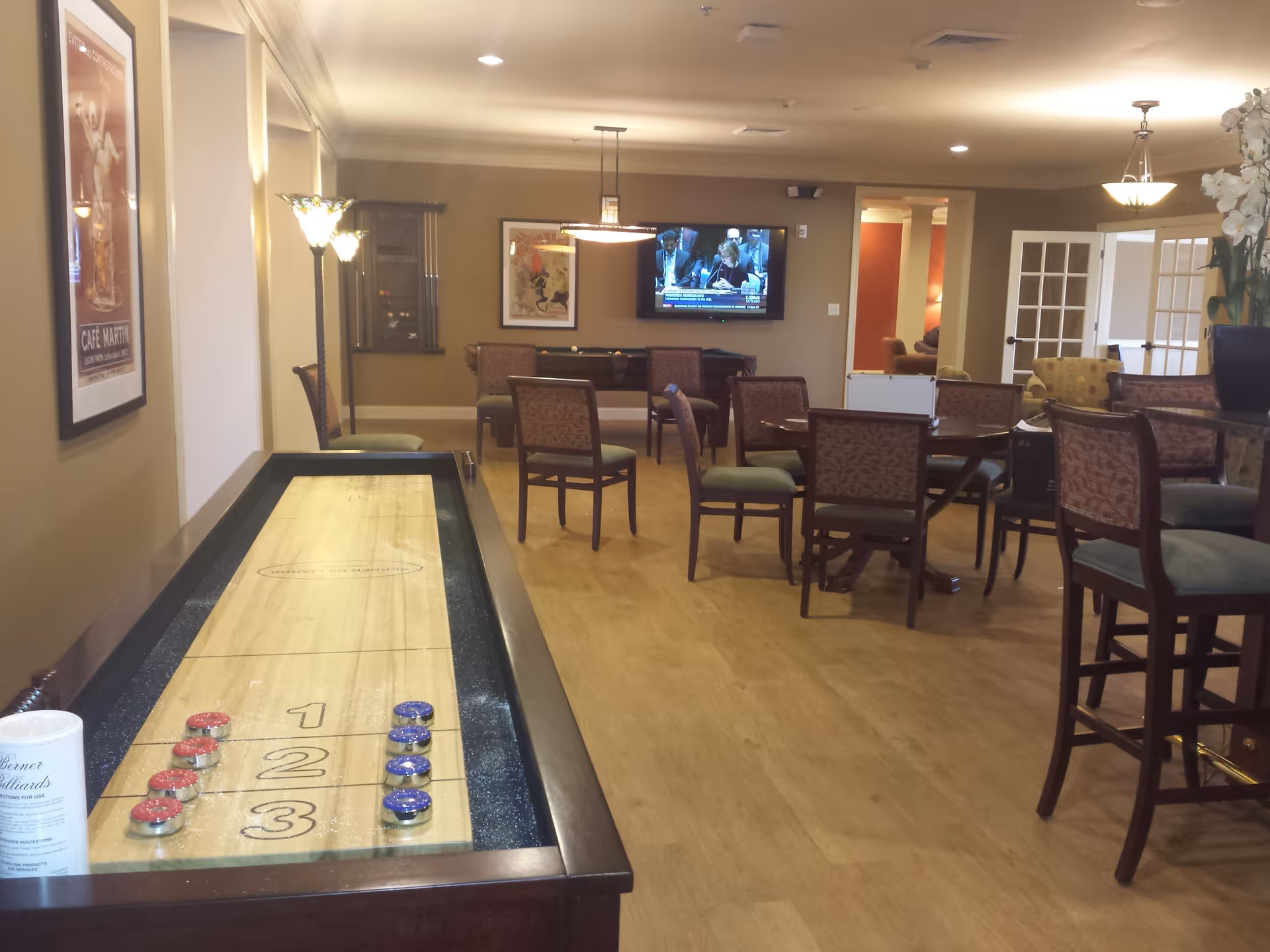 A senior living facility common room with a shuffleboard table in the foreground, several chairs and round tables scattered throughout the room, a wall-mounted TV showing a news program, and framed artwork on the walls. The room has warm lighting and a wooden floor.