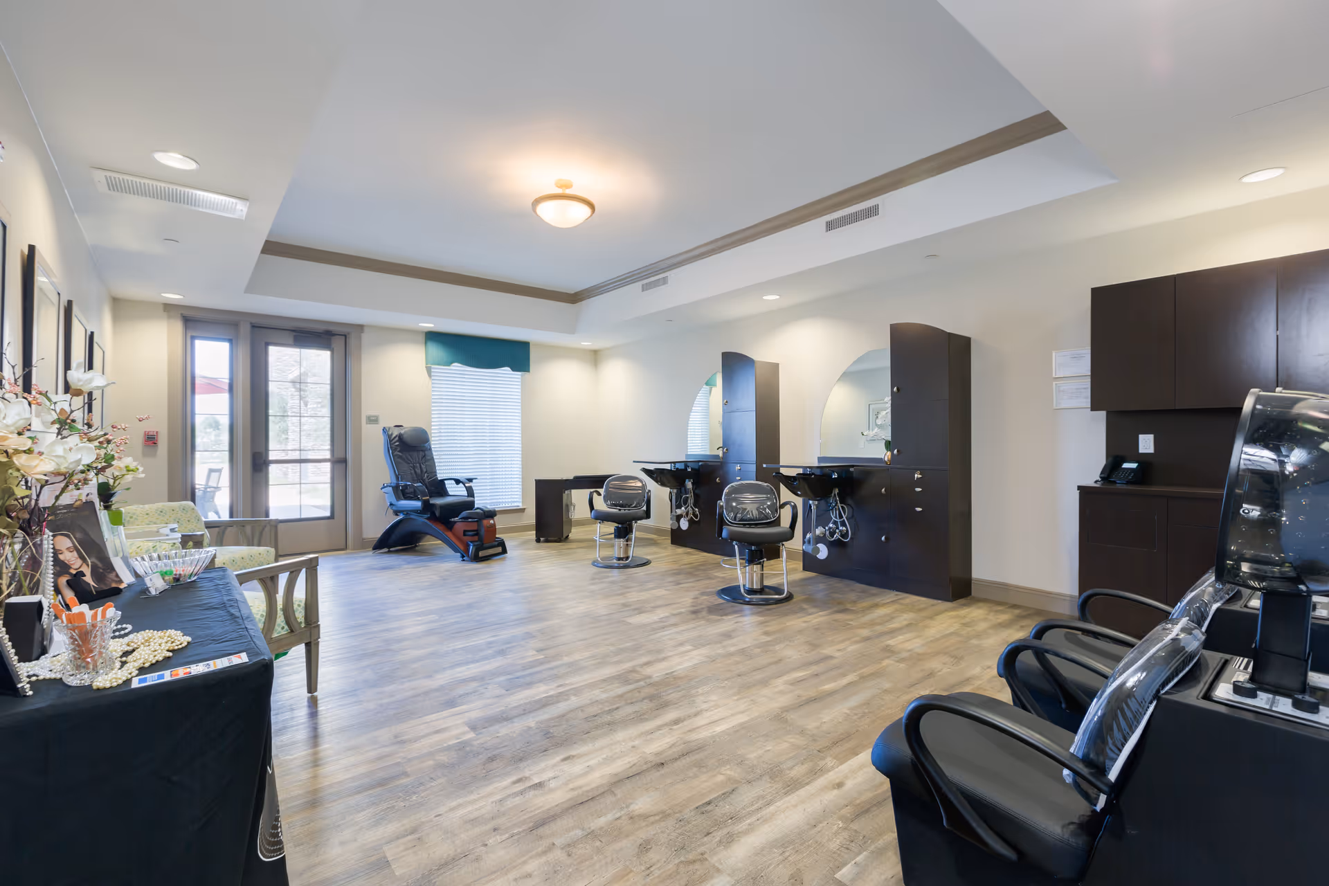 Interior view of a senior living facility's salon area with wooden flooring, two styling chairs in front of mirrors and sinks, a massage chair near a window, and a table with flowers and magazines on the left side.
