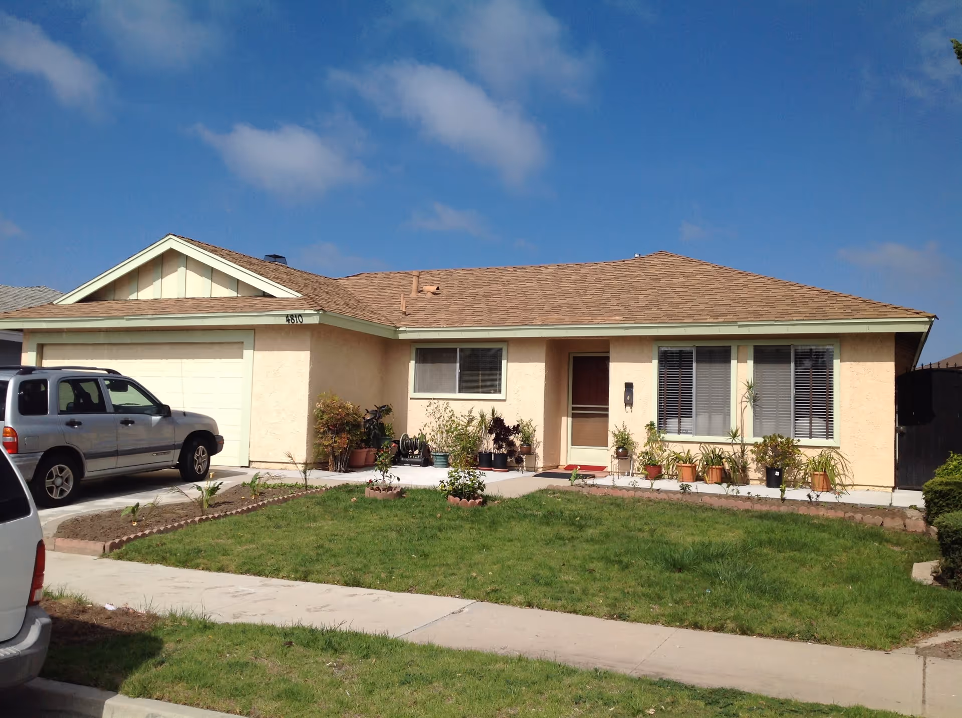 Single-story beige house with a front lawn, driveway and a parked silver SUV under a blue sky.