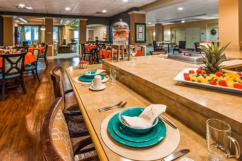 Interior view of a dining area in a senior living facility with tables and chairs set for a meal. The foreground shows a counter with place settings including plates, napkins, cups, and utensils, along with a tray of fresh fruit and a beverage dispenser. The background features more dining tables, chairs, and a well-lit, spacious room with warm tones and wooden flooring.
