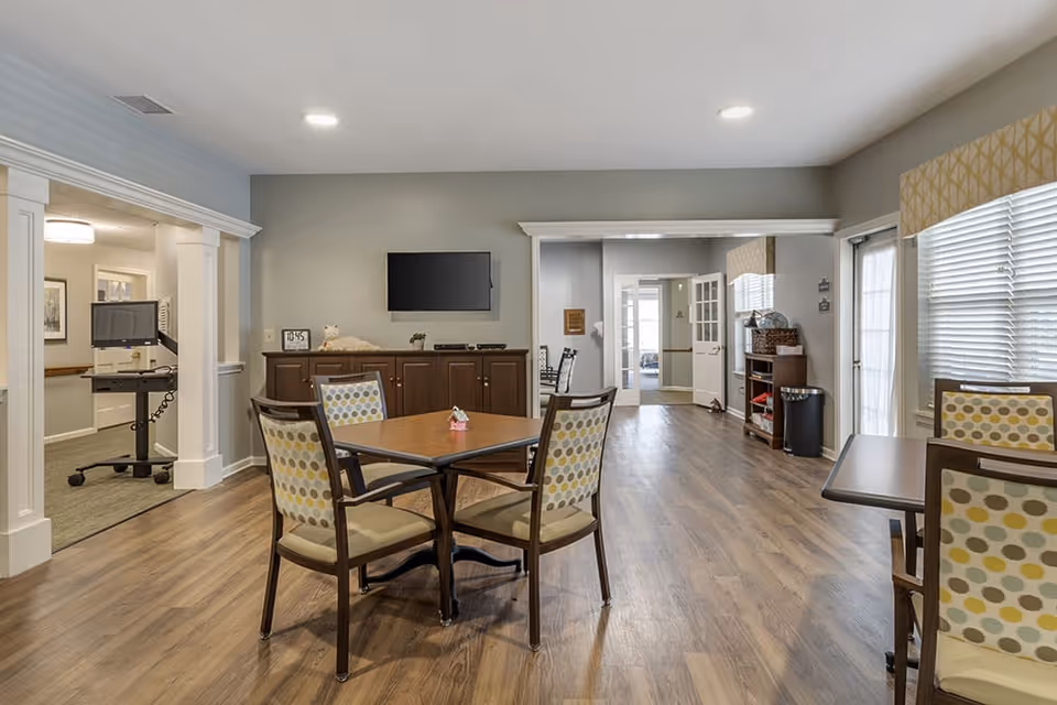 A spacious common area in a senior living facility with wooden flooring, several tables and chairs with patterned upholstery, a wall-mounted flat screen TV, and large windows with blinds allowing natural light to enter. The room has neutral-colored walls and an open doorway leading to other parts of the facility.