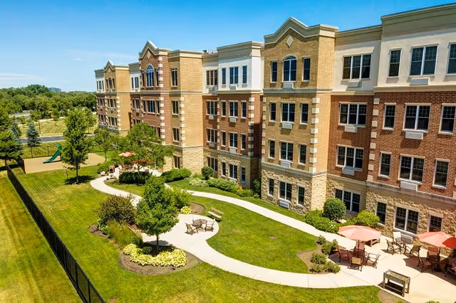 Exterior view of a multi-story assisted living facility with a well-maintained lawn, walking paths, outdoor seating areas with tables and umbrellas, and landscaped greenery under a clear blue sky.