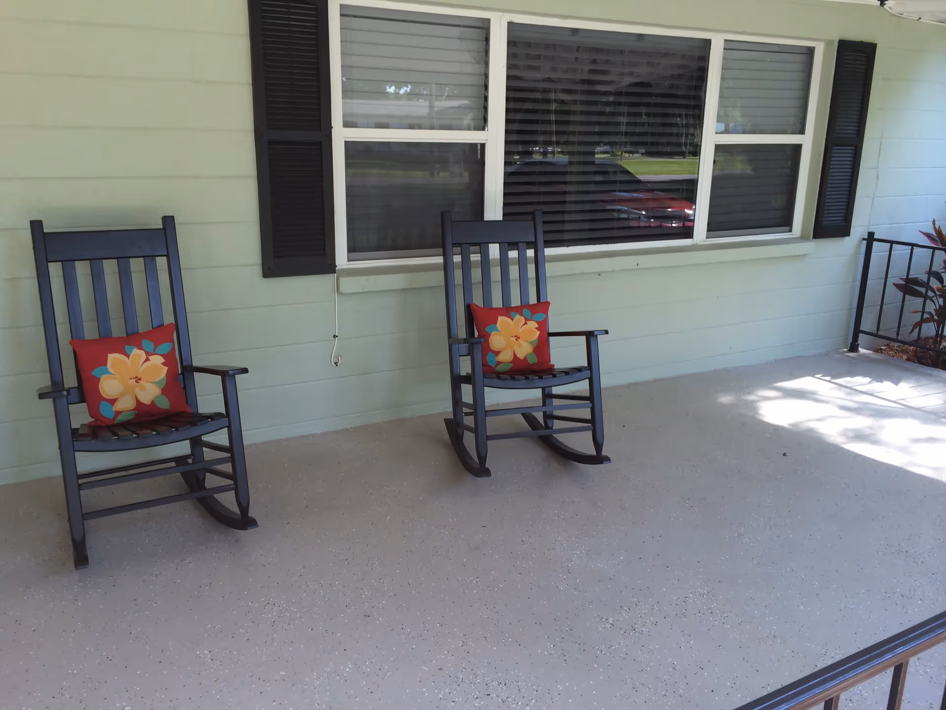Two black wooden rocking chairs with red cushions featuring yellow flowers are placed on a light-colored porch in front of a window with black shutters.