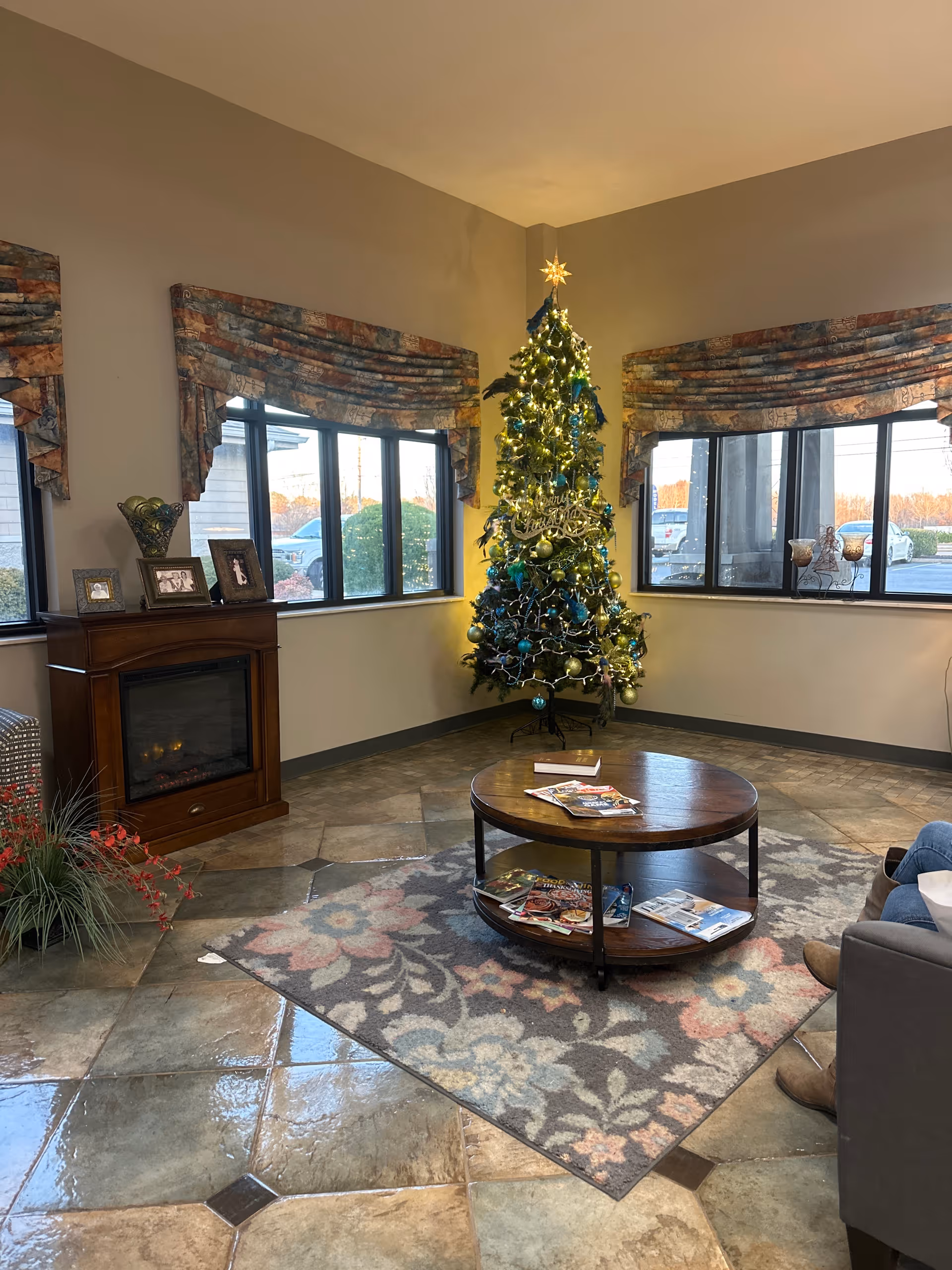 A cozy corner of a living room decorated for Christmas with a lit Christmas tree adorned with ornaments and a star on top. The room has large windows with patterned valances, a wooden electric fireplace with framed photos on top, a round wooden coffee table with magazines, and a floral area rug on a tiled floor. Part of a gray sofa with a person sitting on it is visible on the right side.